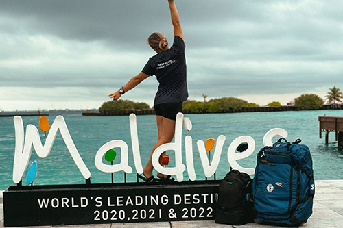 Traveler posing by Maldives destination sign in front of turquoise water with Sport Bag 125 beside them